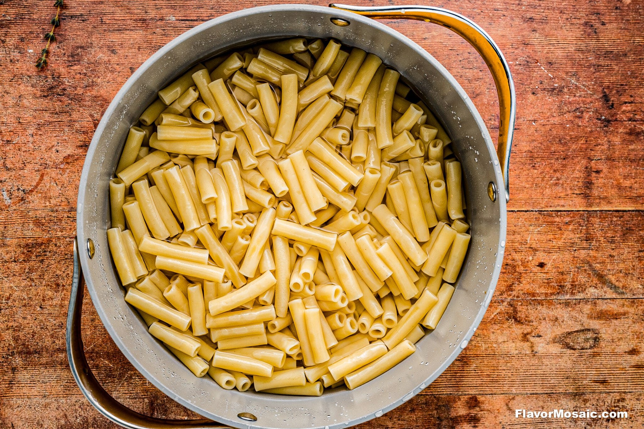 A large metal pot filled with cooked ziti pasta sits on a wooden surface. The pasta is plain, rinsed, and ready for the next step in meal preparation.