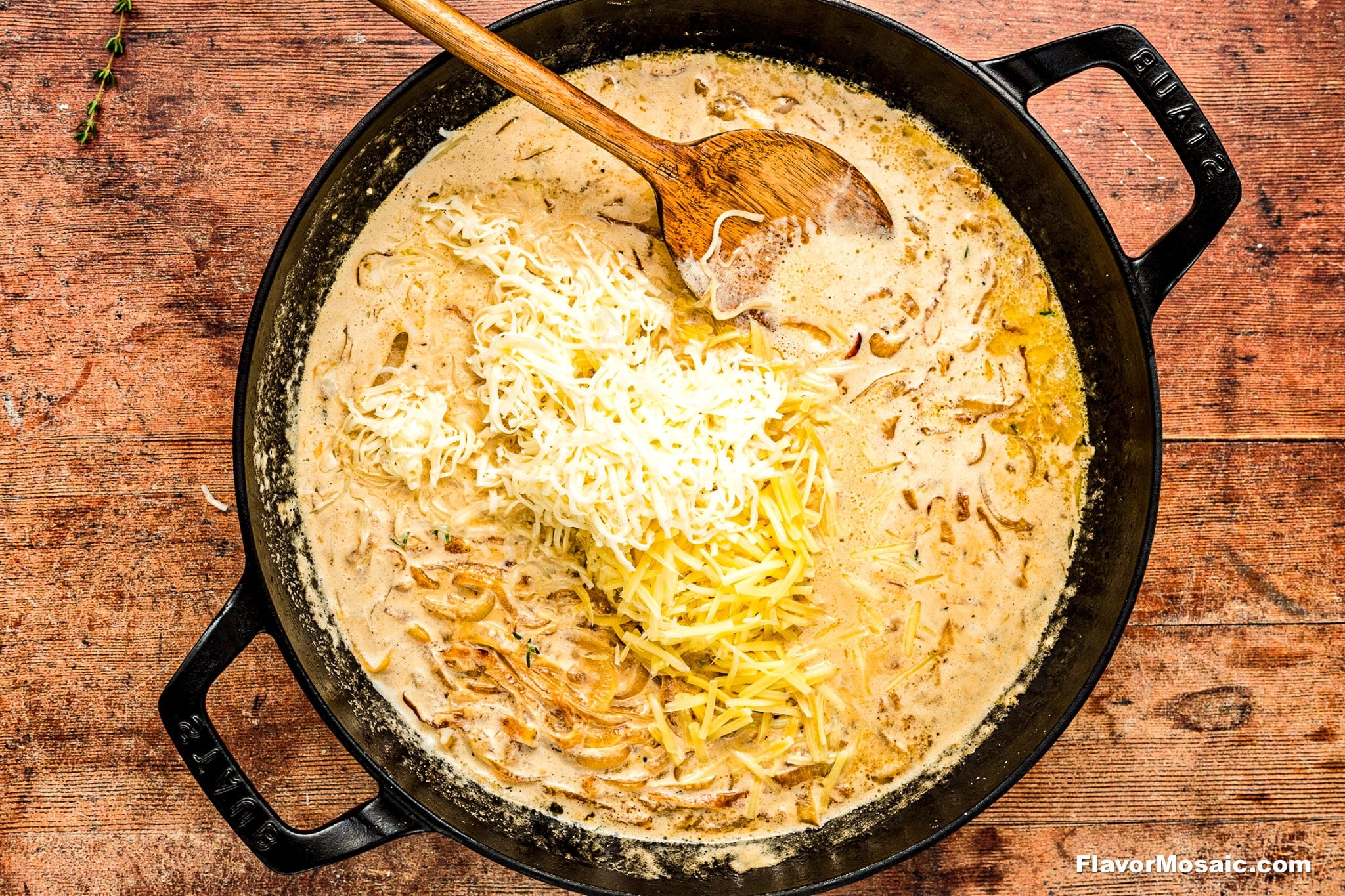 A black cast iron skillet filled with creamy sauce, caramelized onions, and shredded cheese being stirred with a wooden spoon, set on a rustic wooden surface.
