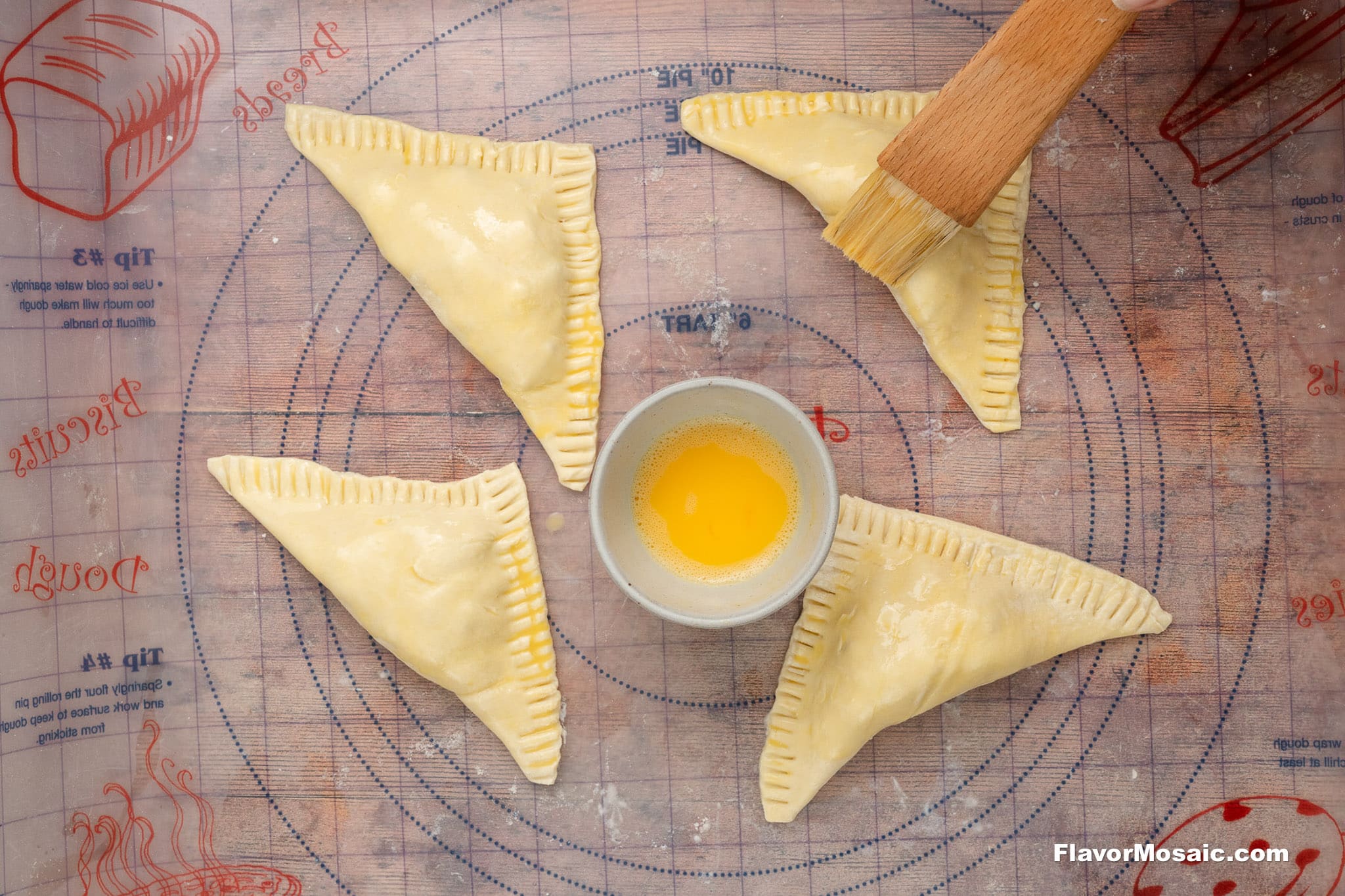 Four uncooked, triangular hand pies with crimped edges are arranged in a circle on a pastry mat. A bowl of egg wash is in the center, and a hand is brushing one pie with a pastry brush.