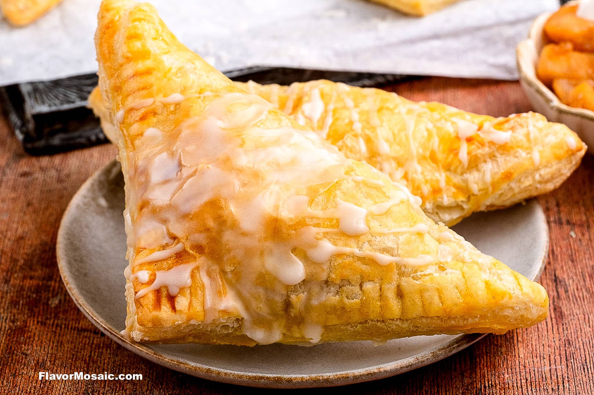 Two golden-brown, triangular apple turnovers with flaky pastry and a drizzle of white icing are served on a plate, with a bowl of diced apples in the background.