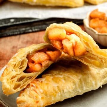 A close-up of a flaky pastry filled with spiced apple chunks, cut in half and stacked on a plate. More pastries and a small bowl of apple filling are visible in the background.