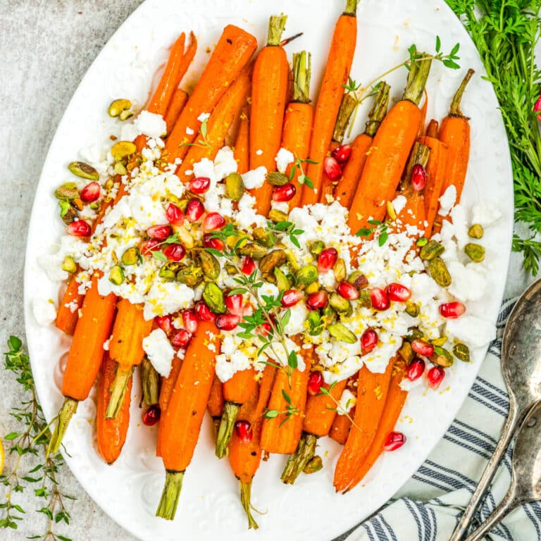 A white oval plate with roasted carrots topped with crumbled feta cheese, pomegranate seeds, pistachios, fresh herbs, and lemon zest, set on a light surface next to a striped towel and serving utensils.