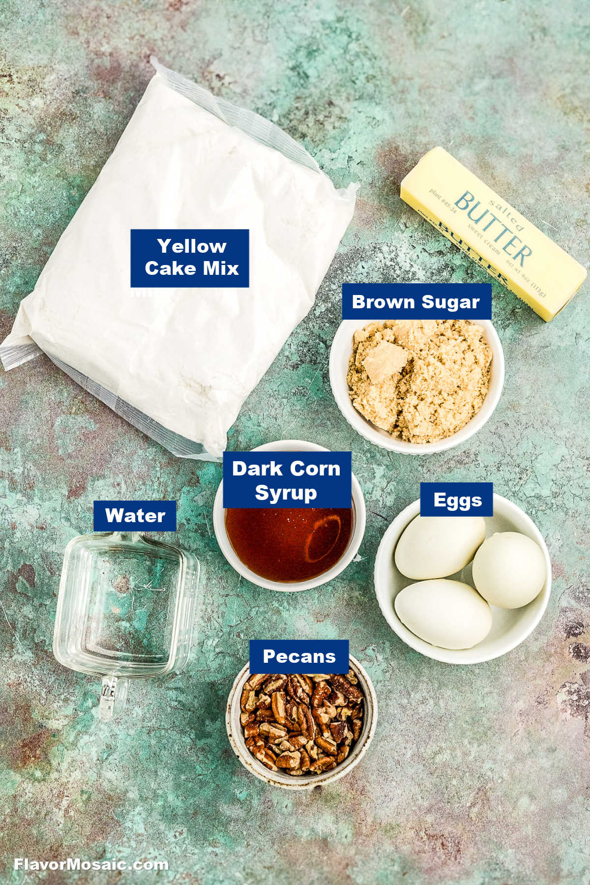A flat lay of ingredients for baking, including a packet of yellow cake mix, a stick of butter, a bowl of brown sugar, a bowl of dark corn syrup, three eggs, chopped pecans, and a measuring cup of water.