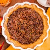 A Texas Pecan Pie in a white pie dish sits on a wooden table, surrounded by autumn leaves, two glasses of orange drink, metal forks on a plate, and a yellow cloth napkin.