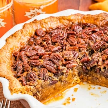 A Texas Pecan Pie in a white pie dish with a slice missing reveals its gooey filling and crunchy pecan topping. Two orange candles with leaf designs and autumn decorations add warmth to the background.