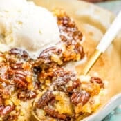 A close-up of pecan pie cobbler served on a plate, topped with a scoop of vanilla ice cream. A spoon in the foreground holds a bite of the cobbler, showing its crumbly texture and pecan pieces.