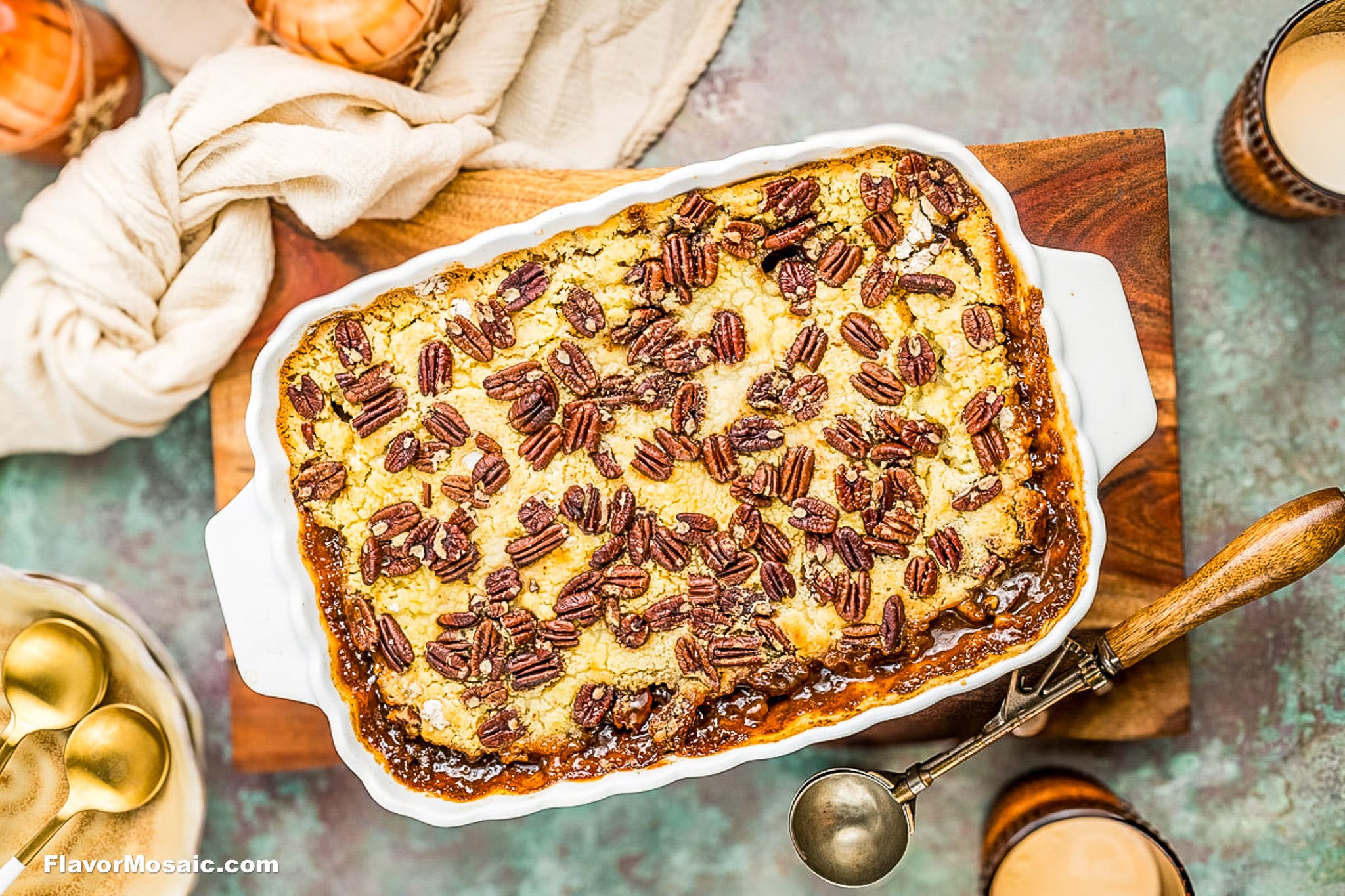A rectangular baked dessert topped with pecans sits in a white dish on a wooden board, surrounded by a serving spoon and drinks. The golden, nutty surface appears gooey and inviting.