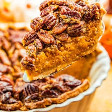 A close-up of a slice of pecan pie being lifted from a pie dish, showing a golden, nutty topping and a rich, moist filling, with autumn-themed decorations blurred in the background.