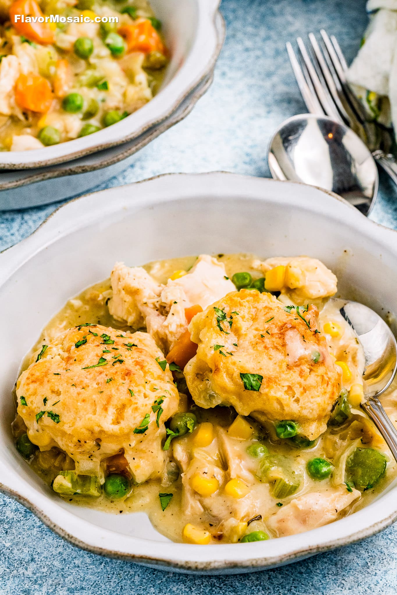 A bowl of Cheddar Bay Biscuit Chicken Pot Pie features creamy chicken and vegetable stew topped with two golden-brown biscuit dumplings, served with a spoon. The hearty mix includes peas, corn, carrots, and chunks of chicken. Another bowl sits in the background.