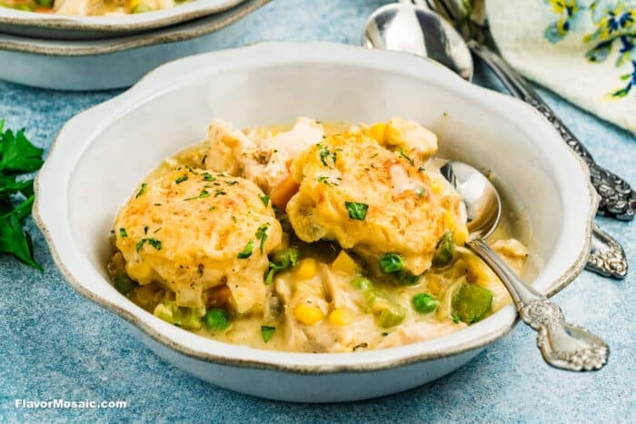 A bowl of chicken and vegetable stew topped with two golden-brown biscuit dumplings, garnished with parsley, with a spoon resting on the side of the bowl.