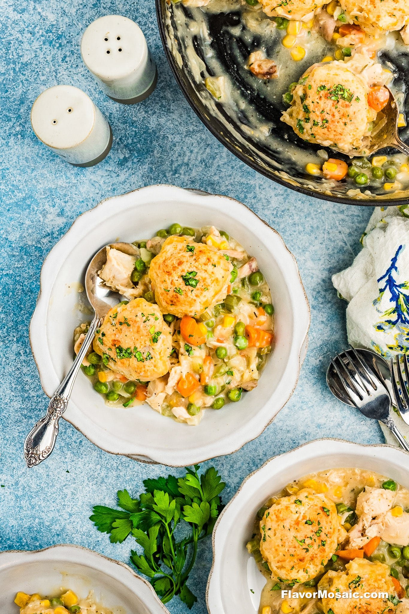 A serving of chicken and vegetable stew topped with golden herb biscuits in white bowls, with a skillet of the same dish, salt and pepper shakers, and fresh parsley on a blue surface.
