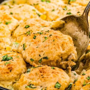 A close-up of a skillet filled with golden-brown biscuit-topped chicken and vegetable pot pie, garnished with chopped herbs. A spoon is lifting a portion of the dish.