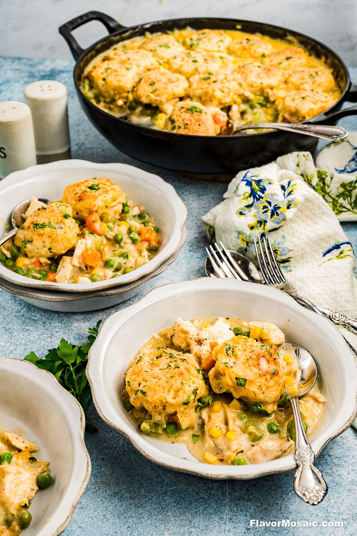 A skillet of Cheddar Bay Biscuit Chicken Pot Pie, filled with chicken and vegetables and topped with golden biscuit dumplings. Served in bowls with spoons, alongside a floral napkin, herbs, and salt and pepper shakers.