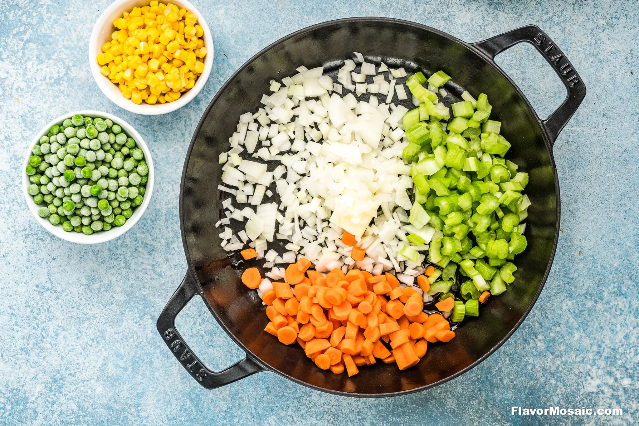 A black pot contains diced onions, celery, and carrots on a blue surface-perfect for starting a Chicken Pot Pie with Cheddar Bay Biscuits. Nearby are small bowls with frozen peas and corn.