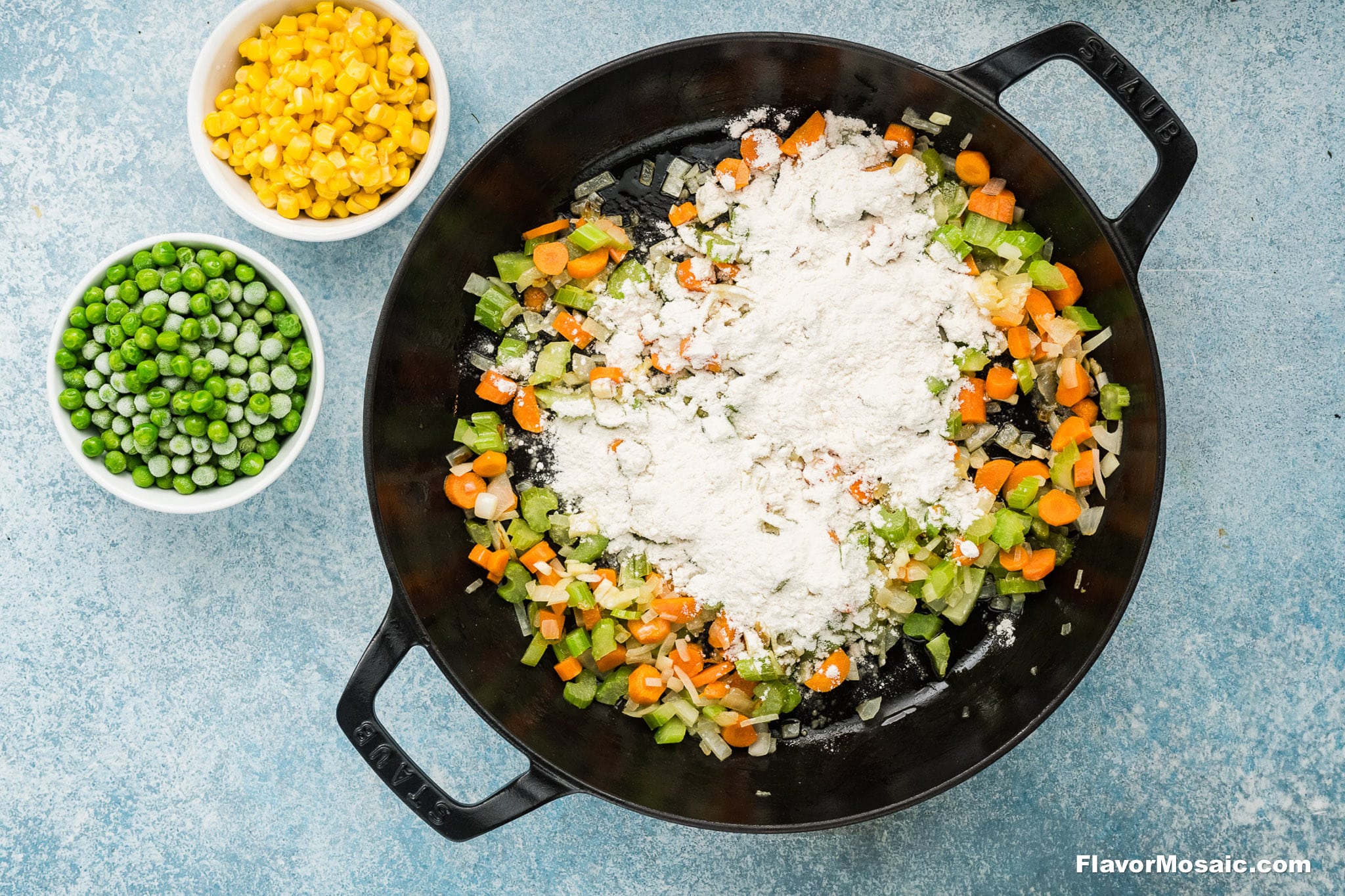 Overhead view of a black skillet with chopped carrots, celery, onions, peas, and flour-perfect ingredients for a Cheddar Bay Biscuit Chicken Pot Pie-next to bowls of frozen peas and corn on a blue surface.