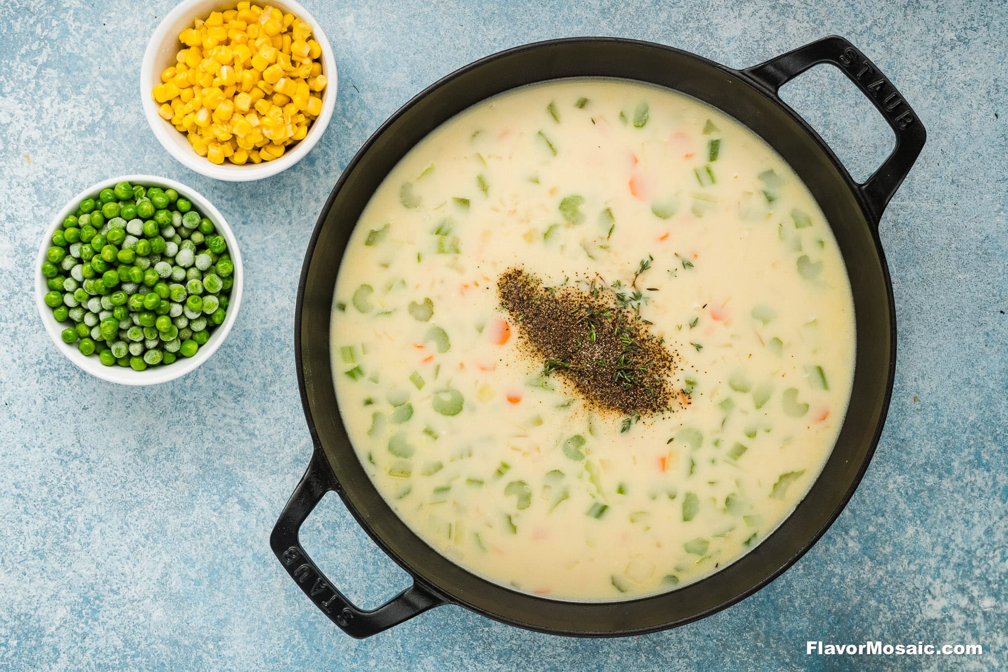 A black pot filled with creamy vegetable soup, reminiscent of Cheddar Bay Biscuit Chicken Pot Pie, is topped with herbs and sits on a light blue surface. Next to the pot are small bowls containing yellow corn and green peas.