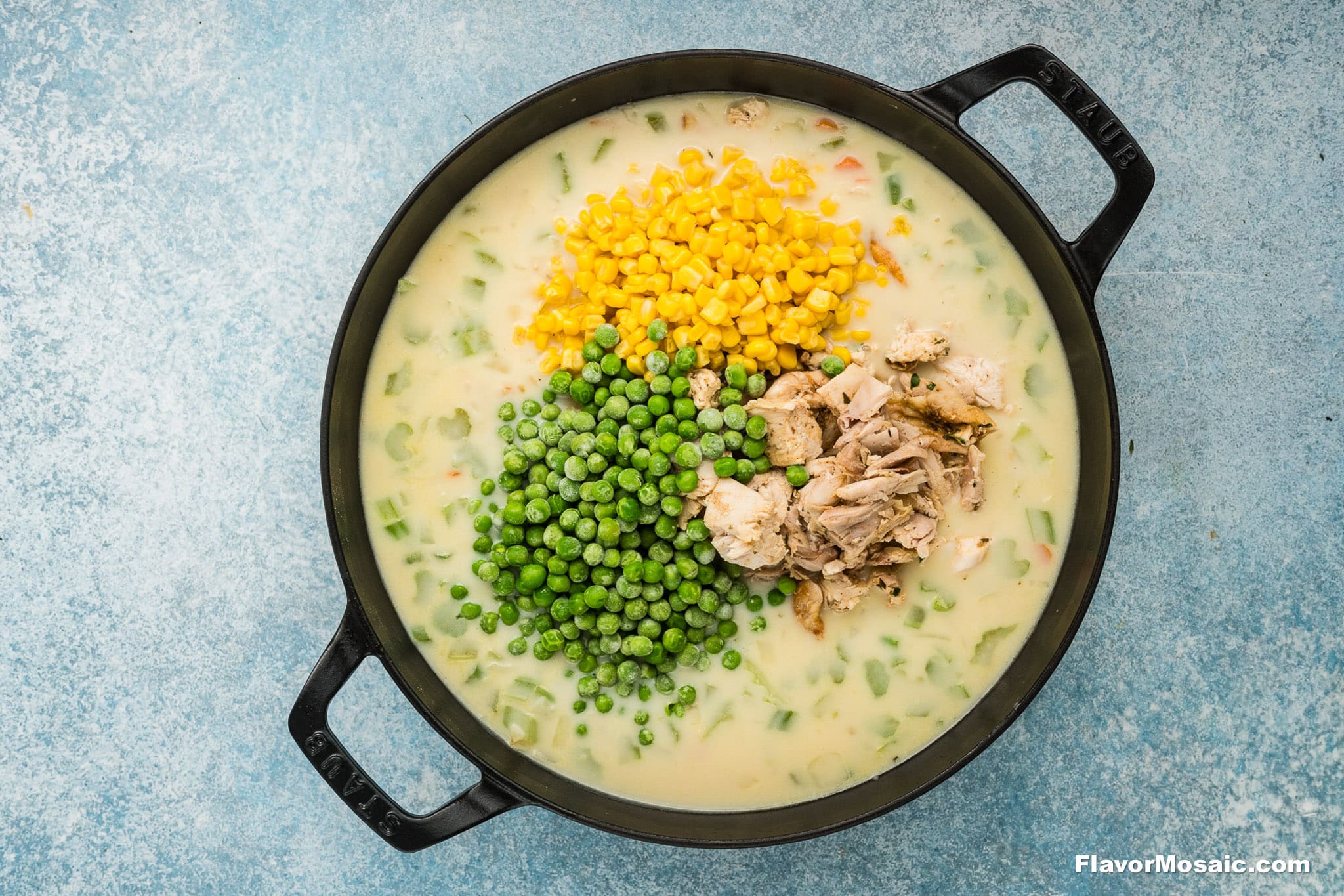 A black pot filled with creamy soup for a Cheddar Bay Biscuit Chicken Pot Pie, topped with piles of yellow corn, green peas, and shredded chicken. The pot sits on a blue textured surface, ready to be mixed and enjoyed.