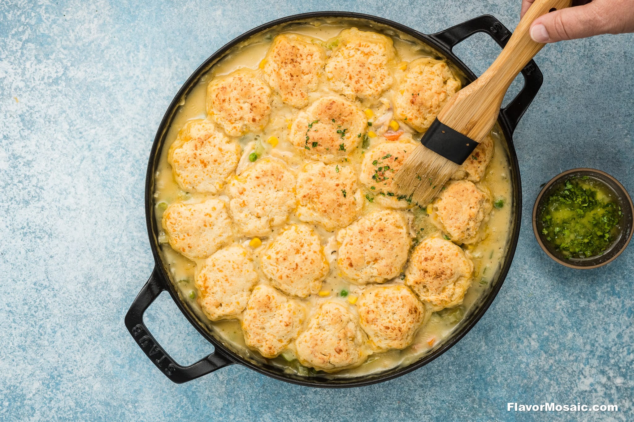 A hand brushes melted butter over golden-brown Cheddar Bay Biscuit topping on a creamy Chicken Pot Pie with Cheddar Bay Biscuits in a black skillet, with a small bowl of herbs on the side.