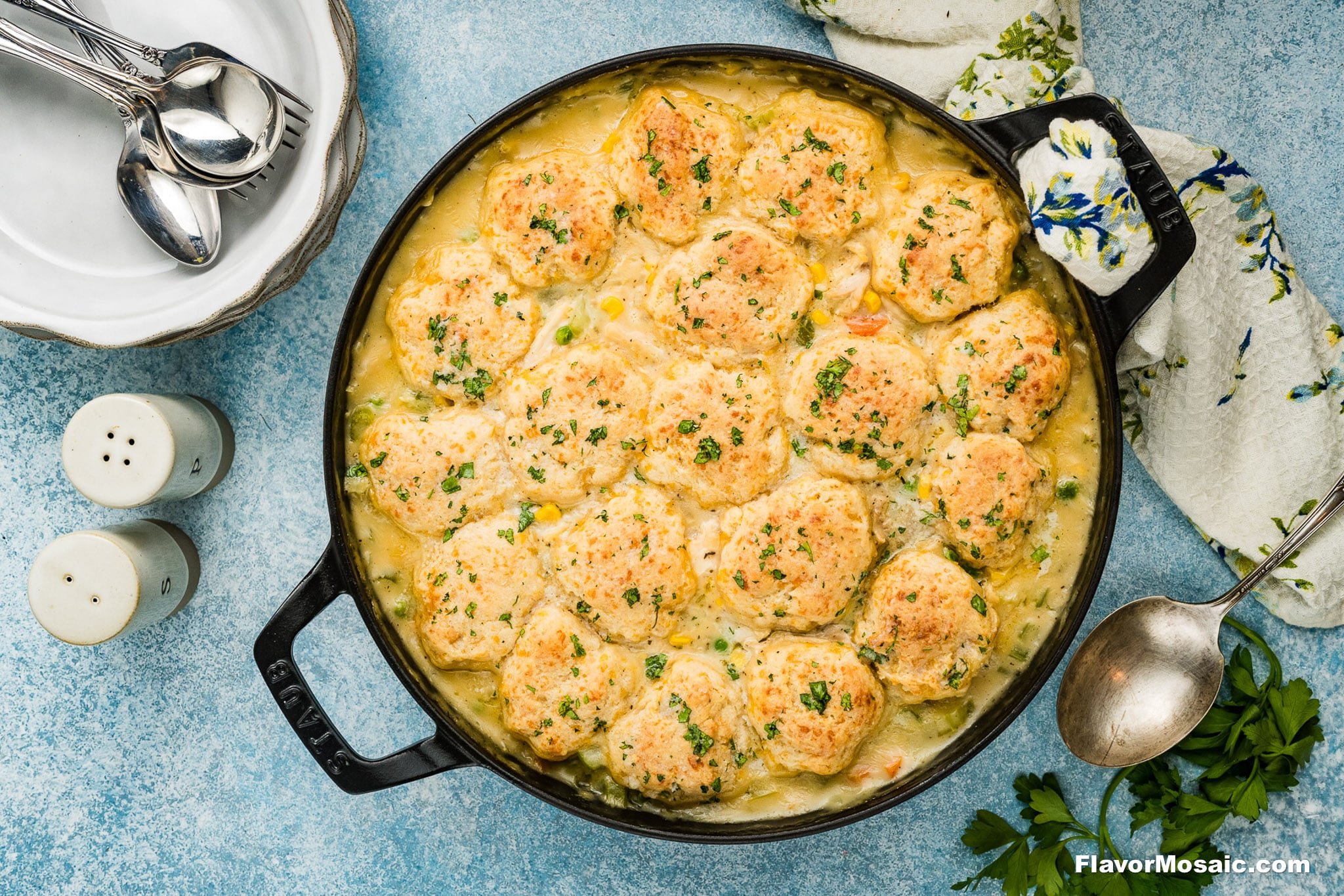 A skillet filled with golden Cheddar Bay Biscuit Chicken Pot Pie on top of a creamy casserole, surrounded by a spoon, stacked plates with spoons, salt and pepper shakers, and a floral napkin on a blue textured surface.