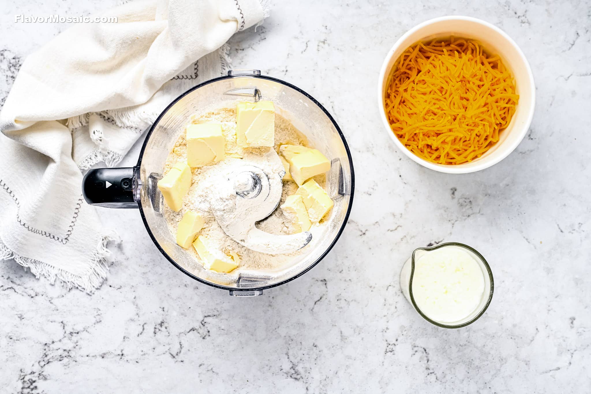 A food processor with flour and butter cubes, a bowl of shredded cheddar cheese, and a measuring cup of liquid, all on a marble countertop with a folded cloth napkin nearby.