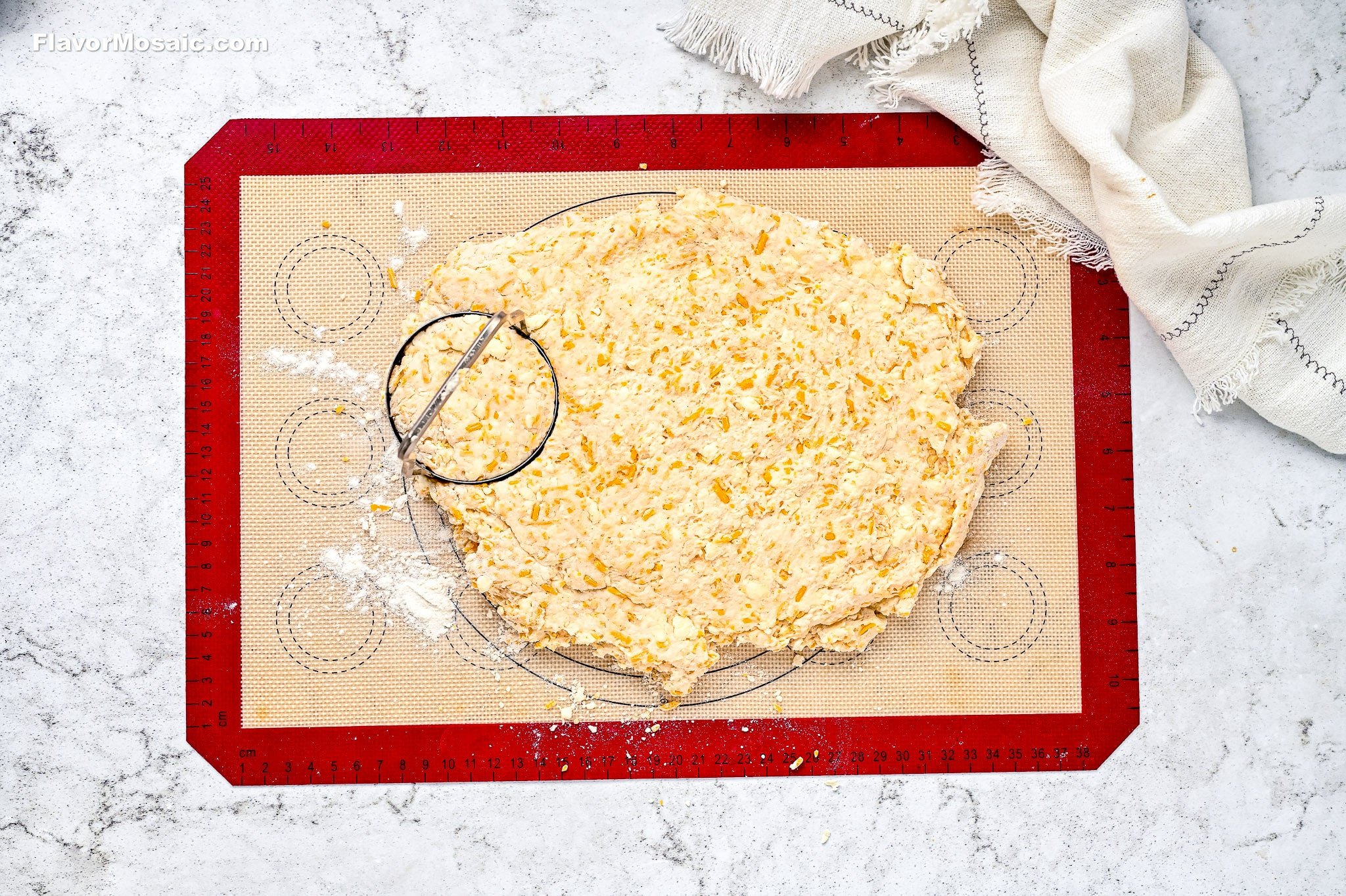 Dough mixed with grated cheese is rolled out on a red silicone baking mat, with a pastry cutter resting on top. A cream-colored towel is in the upper right corner on a marble countertop.