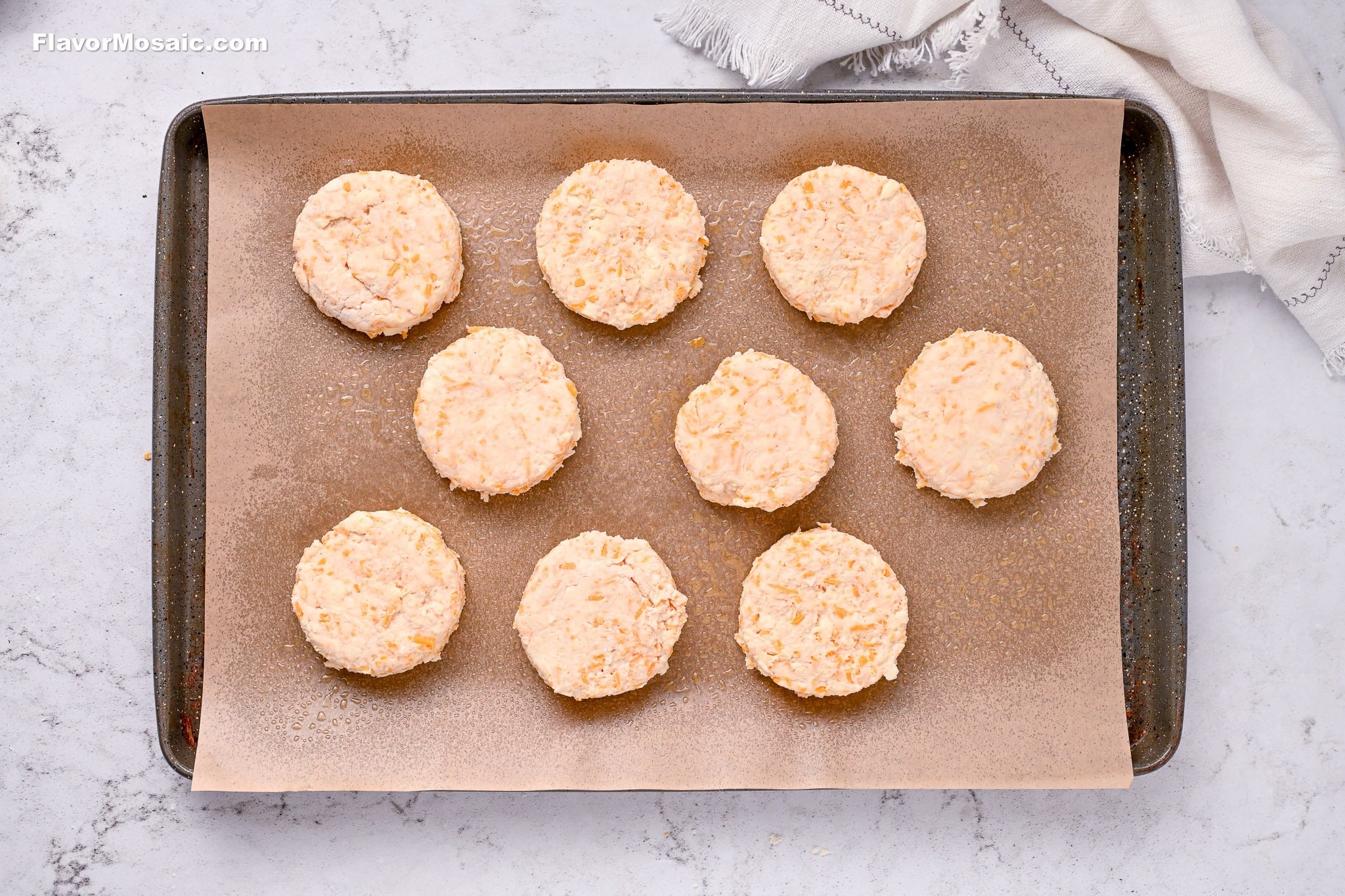 Nine unbaked round biscuits are evenly spaced on a parchment-lined baking sheet, ready to be baked. The tray sits on a light-colored marble surface next to a folded white kitchen towel.