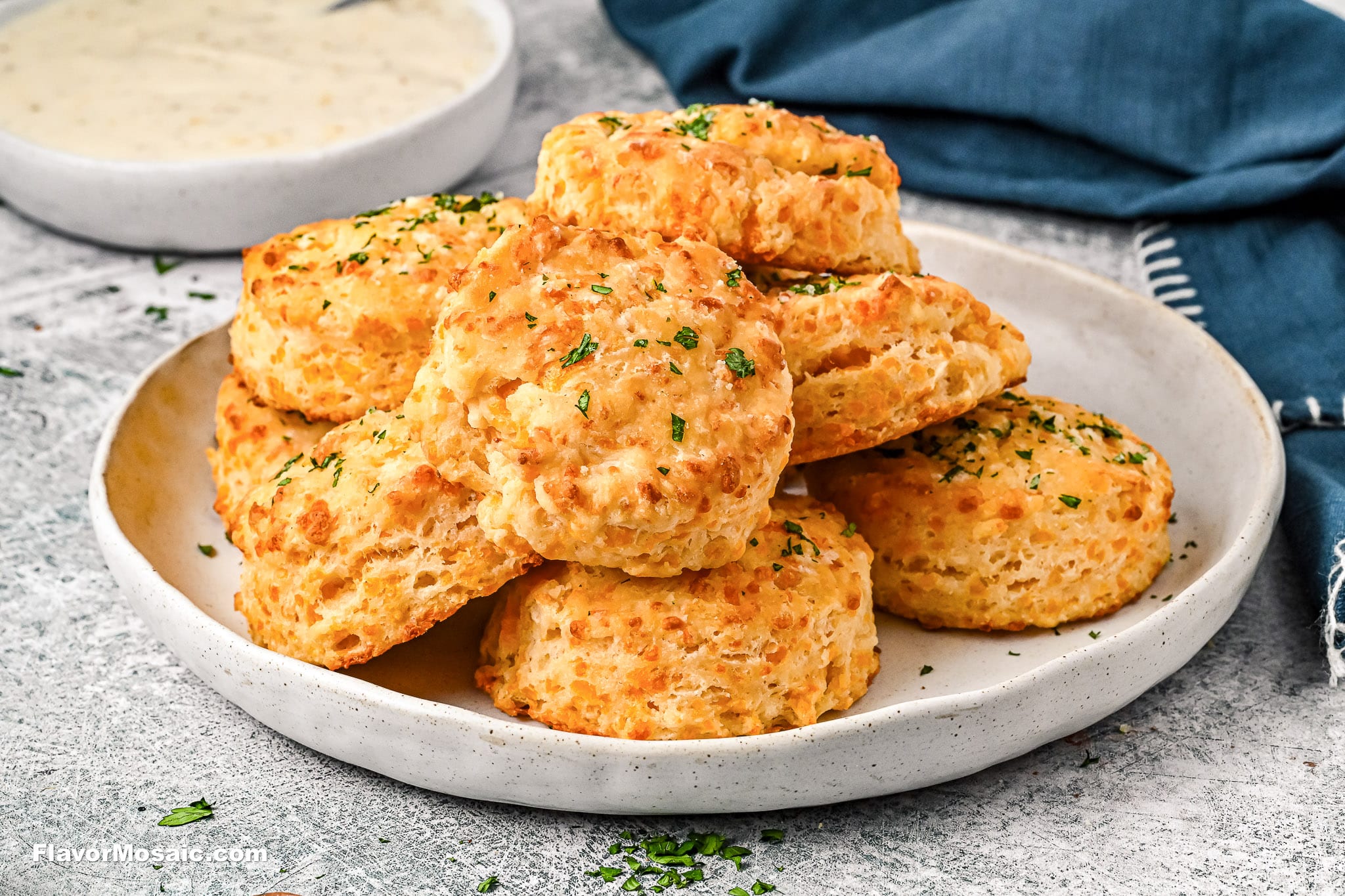 A plate stacked with golden-brown biscuits topped with chopped herbs, with a bowl of creamy dipping sauce and a blue napkin in the background.