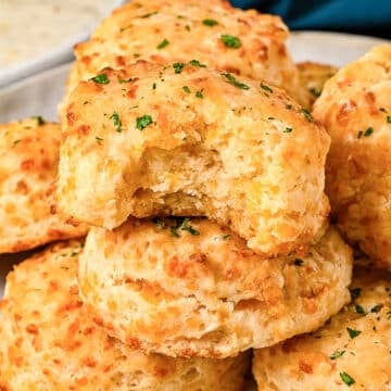 A plate of golden, flaky cheddar biscuits is shown, with one biscuit on top missing a bite. The biscuits are garnished with chopped parsley and look soft and cheesy inside.