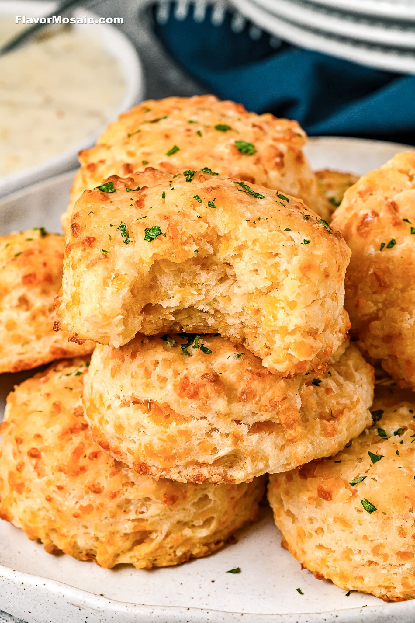 A plate of golden, flaky cheddar biscuits is shown, with one biscuit on top missing a bite. The biscuits are garnished with chopped parsley and look soft and cheesy inside.