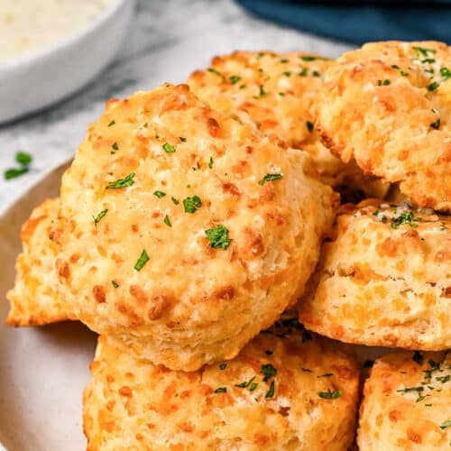 A plate of golden, flaky cheddar biscuits garnished with chopped parsley, with a small bowl of white dipping sauce and a dark blue napkin in the background.