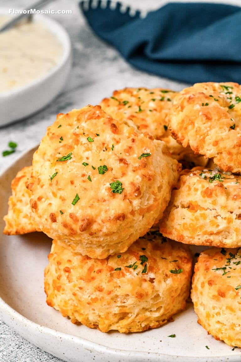 A plate of golden, flaky cheddar biscuits garnished with chopped parsley, with a small bowl of white dipping sauce and a dark blue napkin in the background.