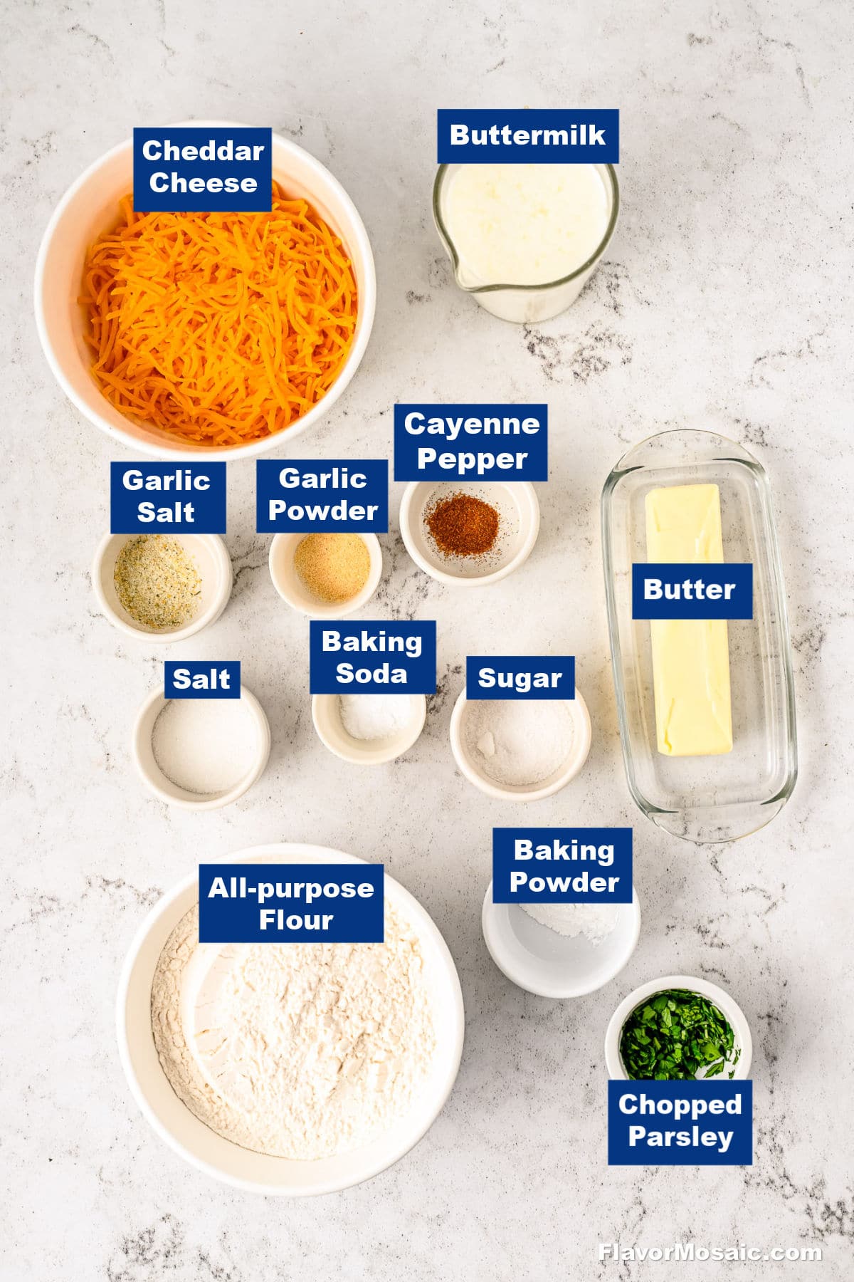 Top-down view of ingredients for a recipe, including cheddar cheese, buttermilk, butter, chopped parsley, all-purpose flour, salt, sugar, cayenne pepper, garlic powder, garlic salt, baking soda, and baking powder in separate bowls.