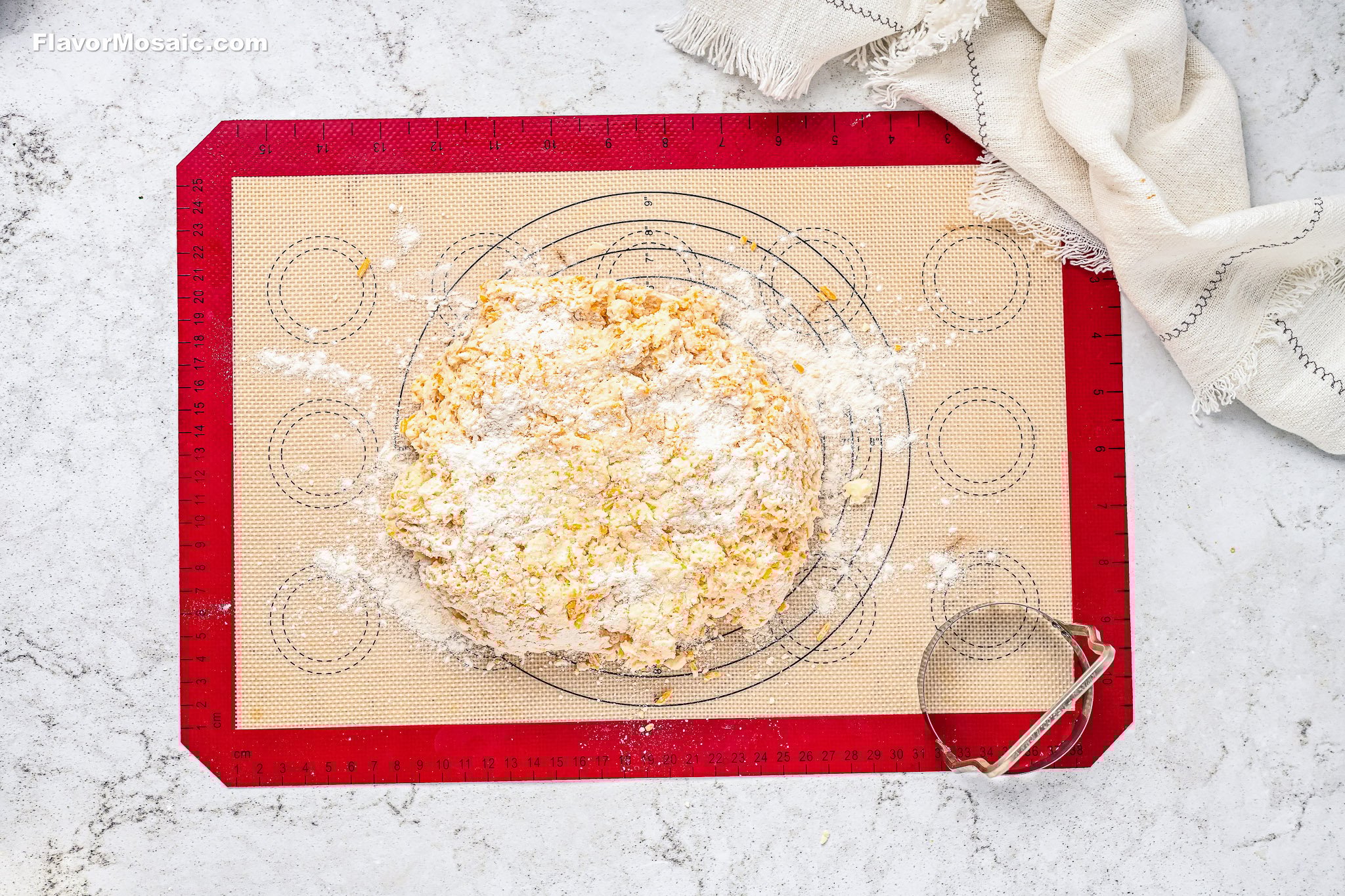 A ball of dough dusted with flour sits on a red and tan silicone baking mat, surrounded by scattered flour. A metal biscuit cutter and a beige cloth are nearby on a white marble surface.