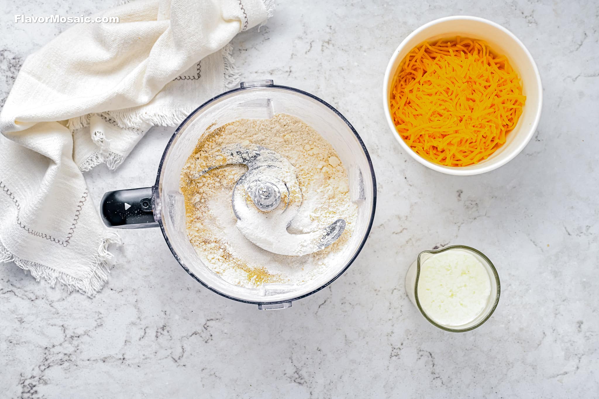 A food processor with flour mixture, a bowl of shredded cheddar cheese, and a measuring cup of milk on a marble countertop with a beige cloth beside them.