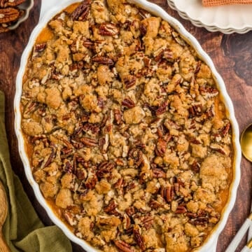 An oval baking dish filled with sweet potato casserole topped with a golden brown pecan and crumb topping, set on a wooden table with plates, napkins, and serving utensils nearby.