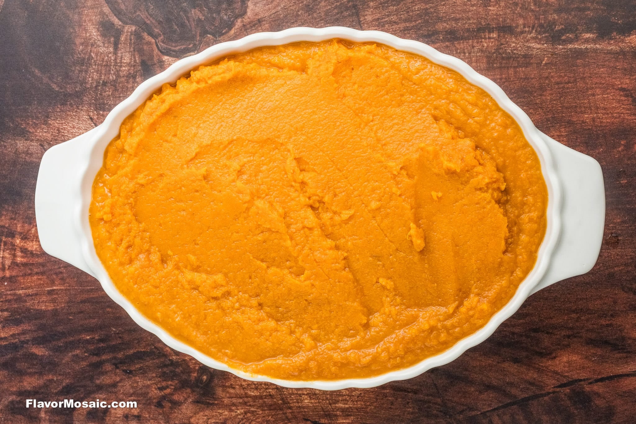 An oval white dish filled with smooth, orange Sweet Potato Casserole with Pecan Topping sits on a dark wooden surface. The casserole has a creamy texture and is evenly spread in the dish.