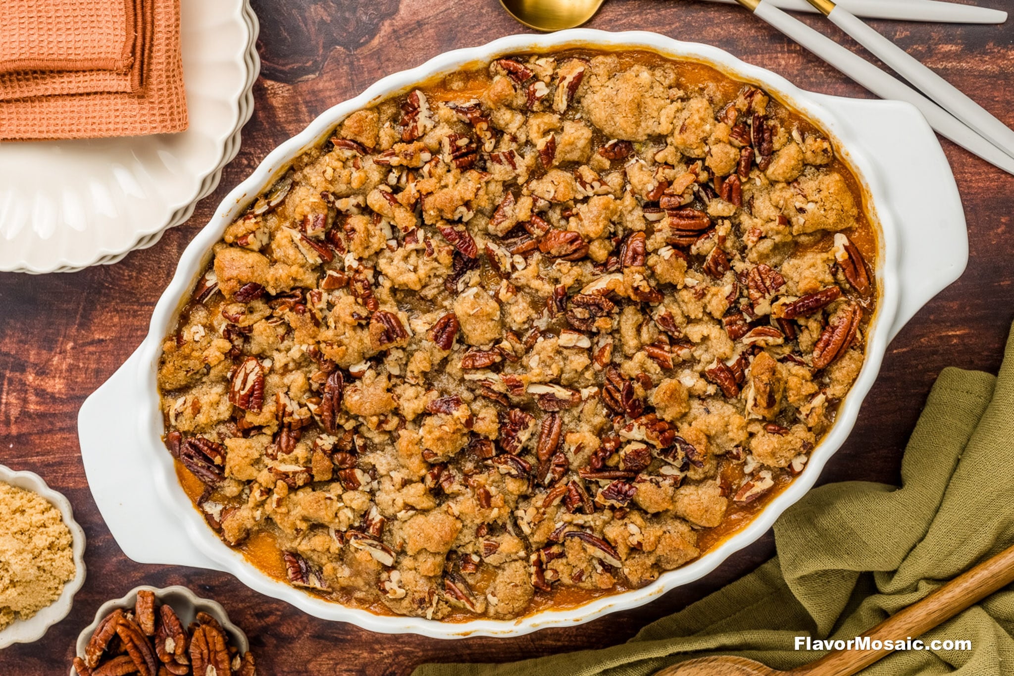 An oval baking dish filled with sweet potato casserole topped with a crumbly brown sugar and pecan streusel, set on a wooden table with plates, utensils, and a folded napkin nearby.