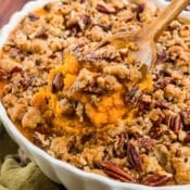 A wooden spoon scoops a portion of sweet potato casserole topped with crunchy pecans and crumbly brown sugar topping from a white scalloped baking dish.