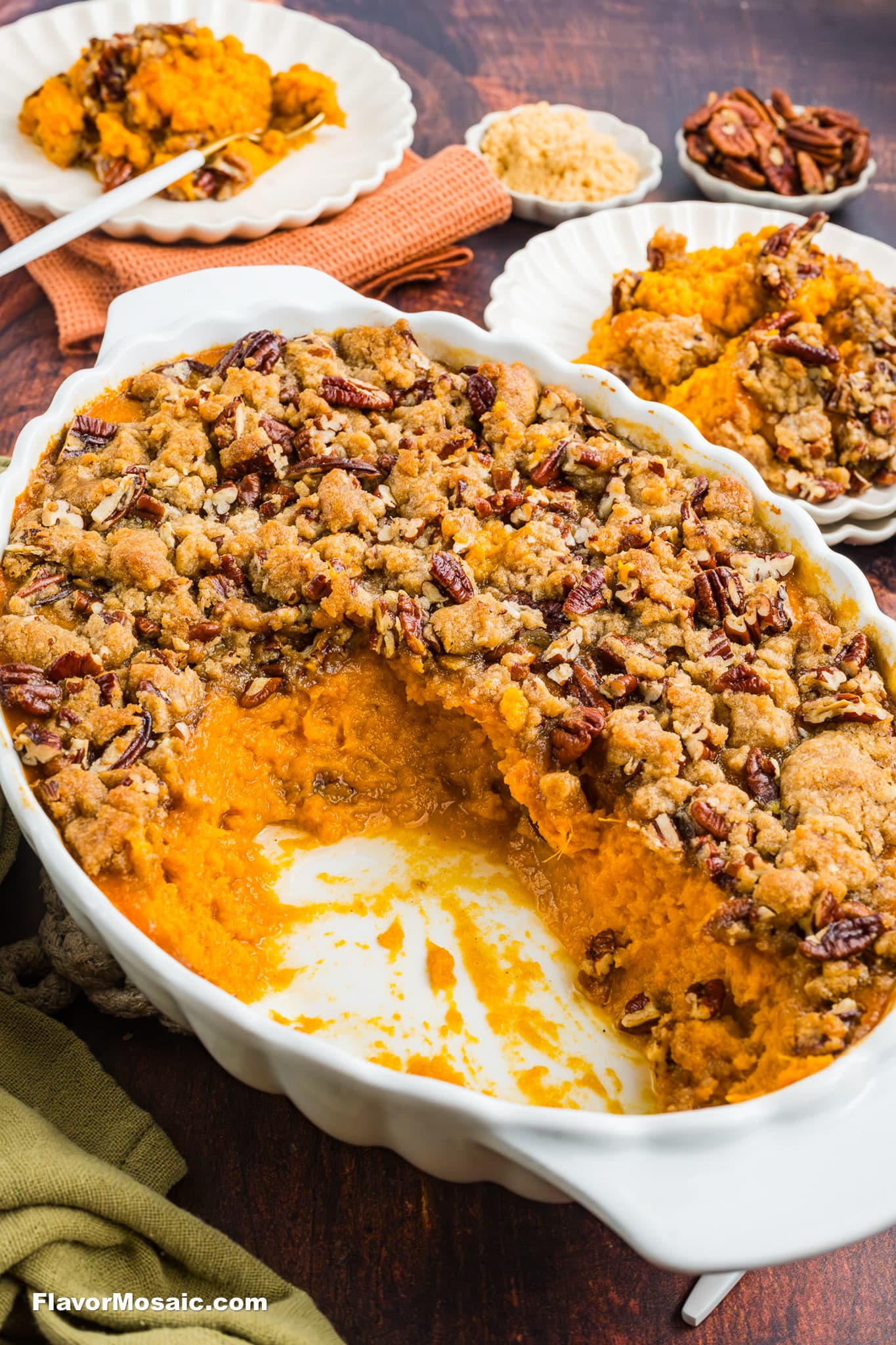 A baked sweet potato casserole with a pecan crumble topping sits in a white dish, with a large portion served out. Plates of casserole and brown sugar are in the background on a wooden table.
