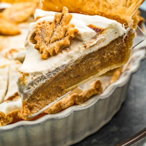 A slice of creamy pumpkin pie with a golden, flaky crust and a dollop of whipped cream, topped with a decorative pastry leaf, being lifted from a ceramic pie dish.