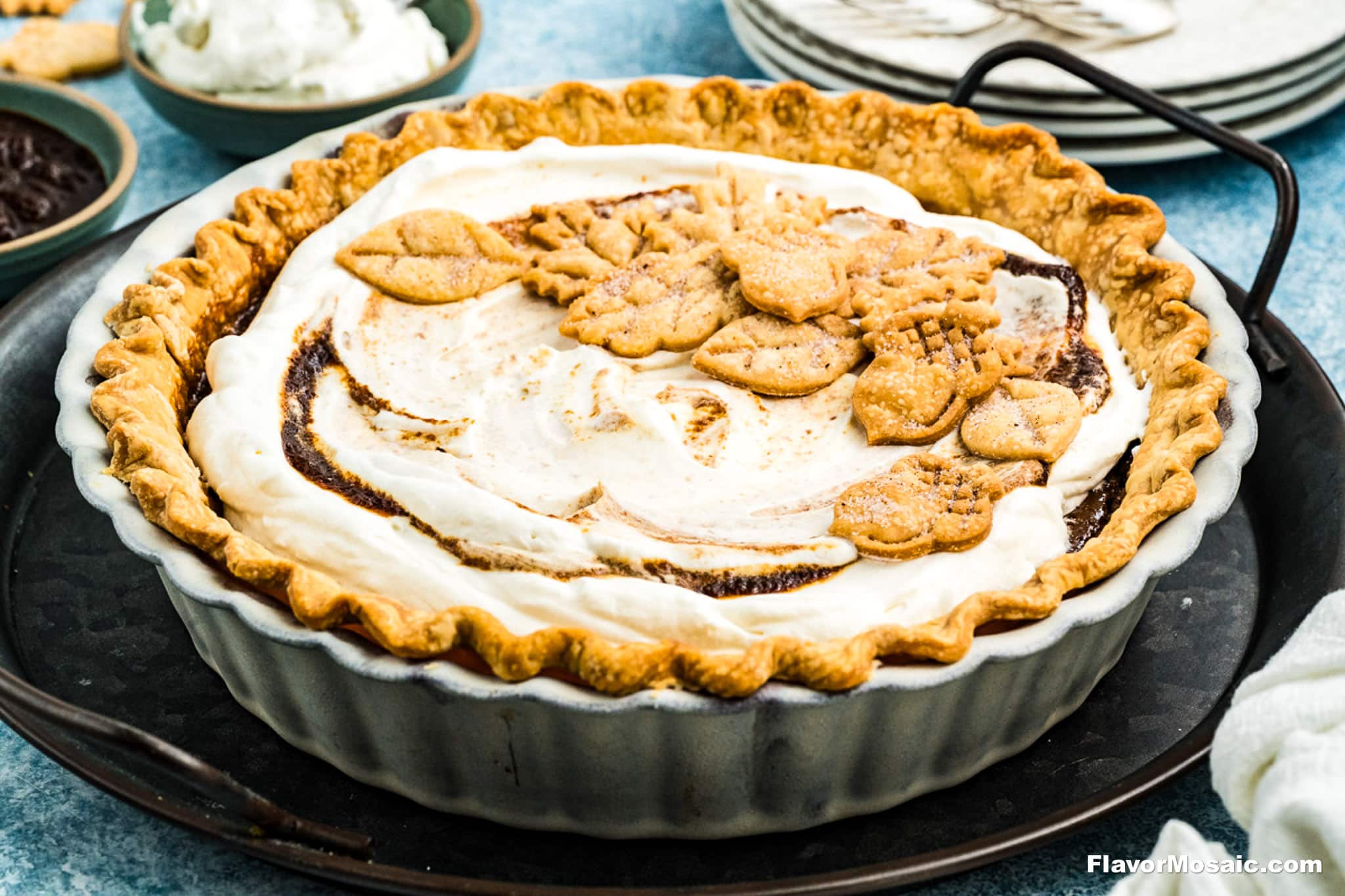 An Apple Butter Pie with a golden, crimped crust, topped with leaf-shaped pastry decorations and swirled cream, sits in a fluted ceramic dish on a tray. Plates and bowls are in the background.