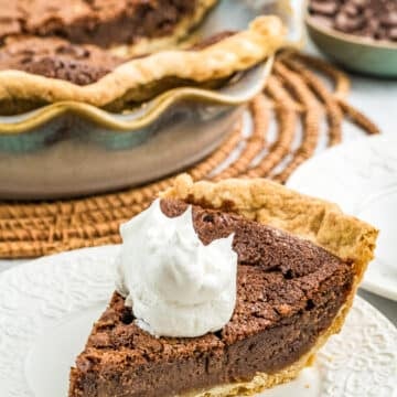 A slice of Chocolate Chess Pie topped with whipped cream sits on a white plate, with the whole pie and chocolate chips in the background.