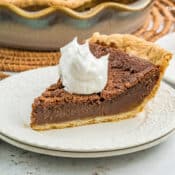 A slice of Chocolate Chess Pie with a flaky crust, topped with a dollop of whipped cream, sits on a white plate. A green striped napkin lies nearby, with another pie in the background.