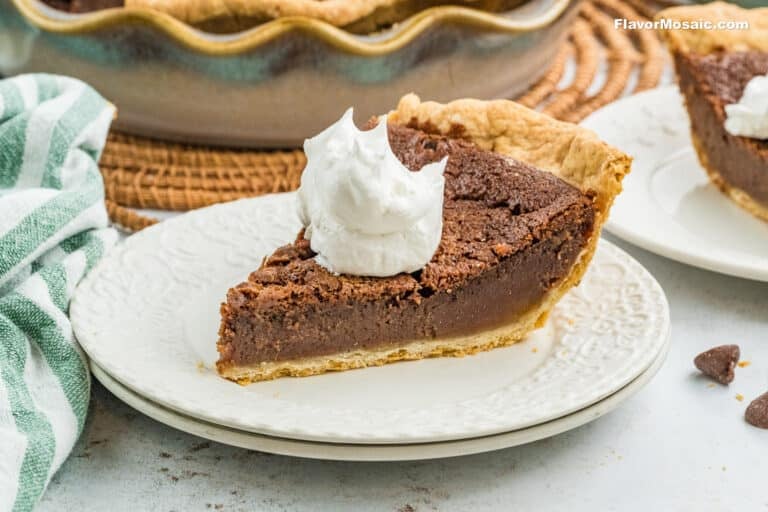 A slice of Chocolate Chess Pie with a golden crust is topped with a dollop of whipped cream, served on a white plate. Another pie and a green-striped towel are visible in the background.
