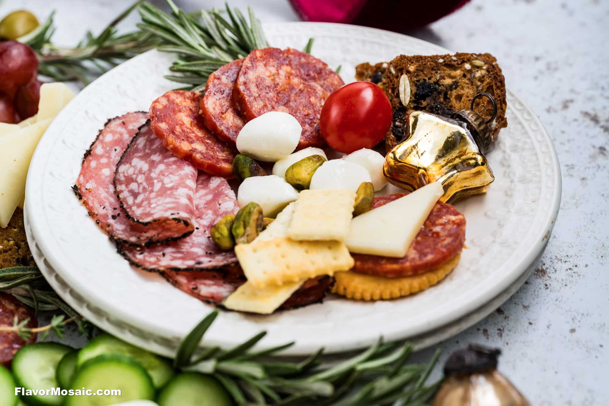 A white plate with assorted charcuterie: sliced salami, pepperoni, cheese, mozzarella balls, pickles, a cherry tomato, crackers, bread, and a shiny gold ornament, garnished with rosemary sprigs.