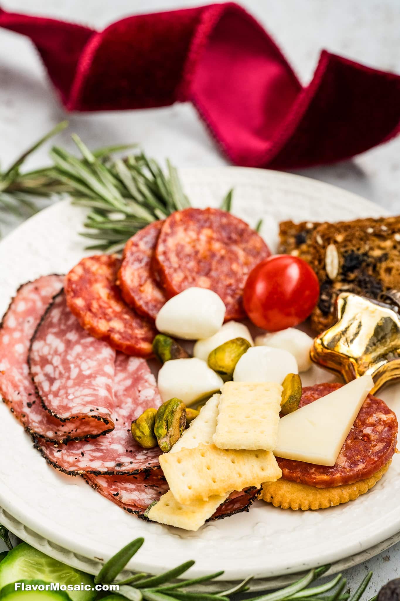 A white plate with sliced salami, pepperoni, crackers, cheese, mozzarella balls, pistachios, a cherry tomato, dark bread, rosemary, and a gold ornament. A red ribbon is in the background.