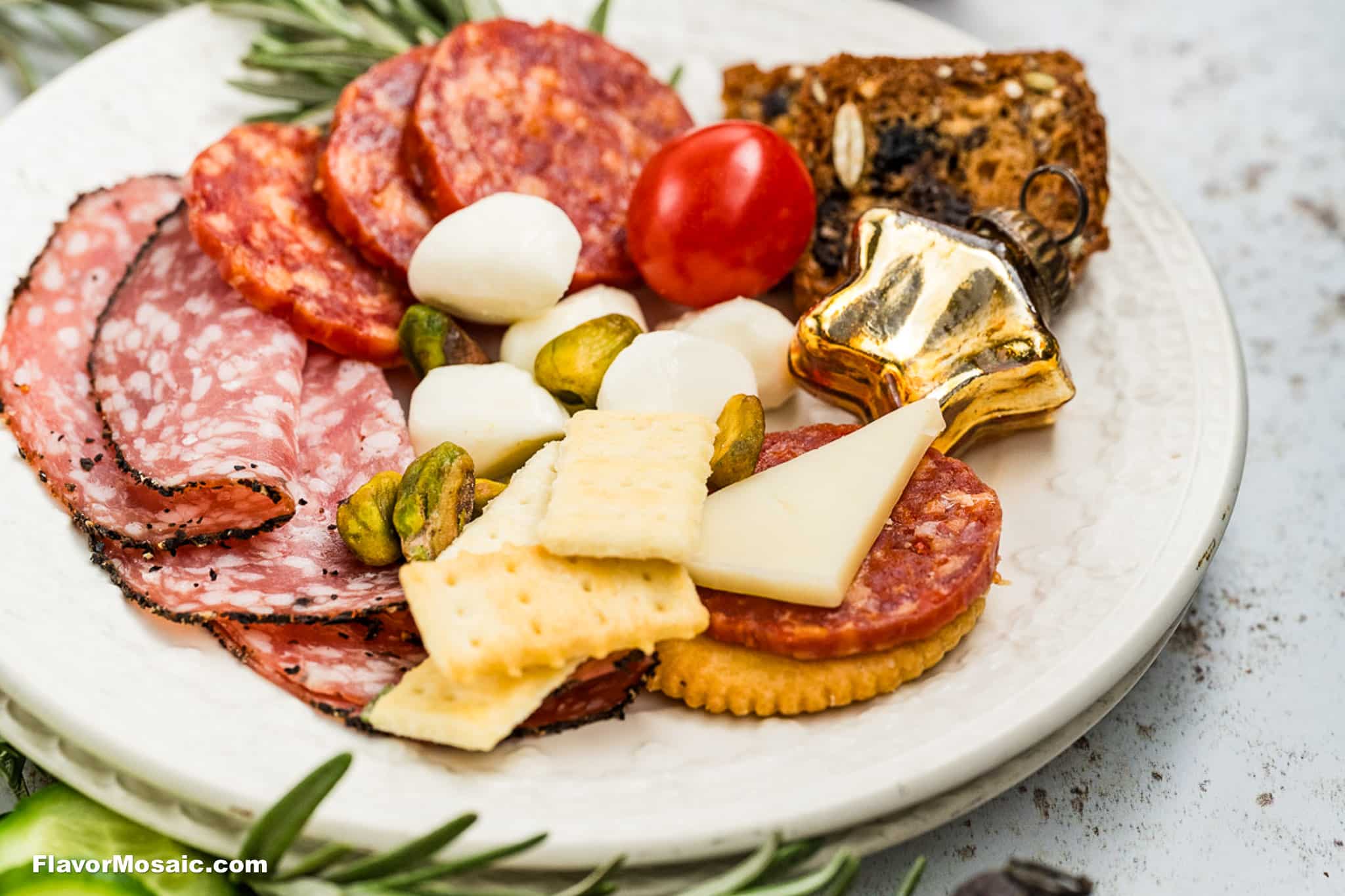 A white plate with salami, pepperoni, mozzarella balls, a cherry tomato, pistachios, crackers, a slice of cheese, a cookie, and a gold ornament, garnished with rosemary.