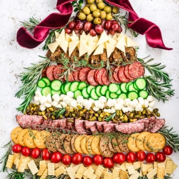 A festive charcuterie board arranged in the shape of a Christmas tree with olives, cheese, meats, cucumbers, tomatoes, crackers, pretzels, and rosemary sprigs, topped with a large red ribbon.