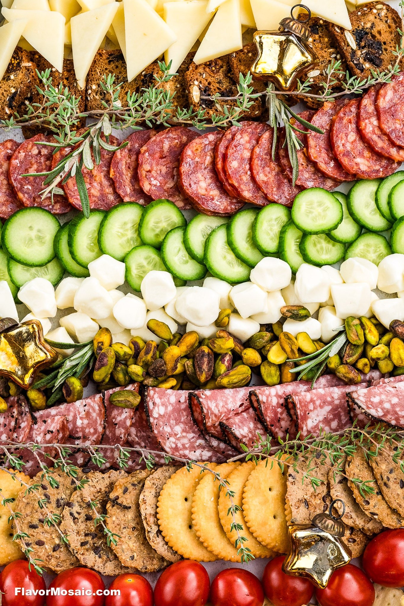 A charcuterie board with rows of sliced cheese, salami, cucumbers, mozzarella balls, pistachios, crackers, and cherry tomatoes, garnished with herbs and decorative gold stars.