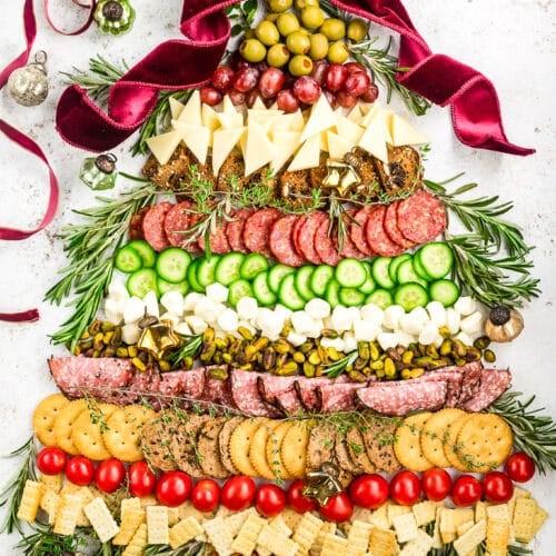 A festive charcuterie board arranged like a Christmas tree with layers of meats, cheeses, crackers, olives, tomatoes, and herbs, topped with a red velvet ribbon bow and Christmas ornaments around it.